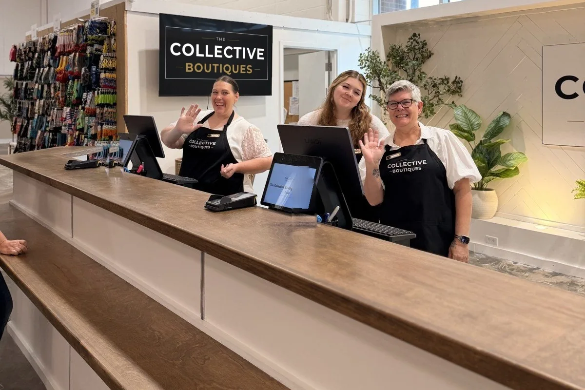 Three staff members waving from the checkout counter at The Collective Boutiques in Overland Park, with Landi CX20 registers, customer-facing displays and Dejavoo payment terminals